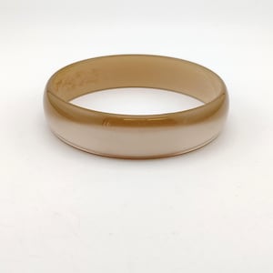 May include: A wide, round bangle bracelet in shades of tan and cream. The bracelet has a smooth, glossy finish and appears to be made of a solid material. The color gradient transitions from a darker tan at the top to a lighter cream at the bottom.