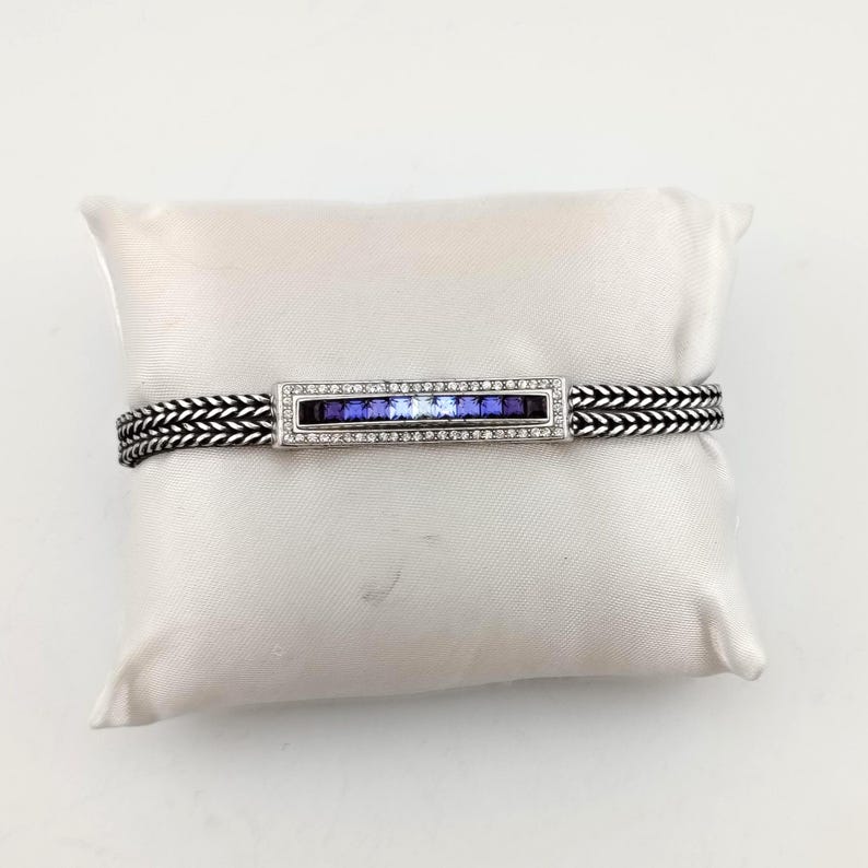 K&ouml;nnte beinhalten: Ein silbernes Armband mit einem rechteckigen Mittelst&uuml;ck mit einem Farbverlauf aus blauen Edelsteinen. Das Mittelst&uuml;ck ist mit kleinen, klaren Steinen eingefasst. Das Armband ist ein gewebtes Design in Schwarz und Silber.