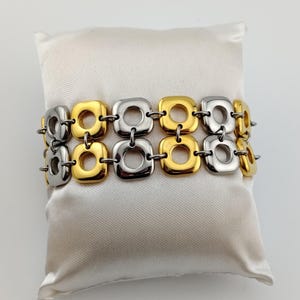 May include: A two-row bracelet with alternating gold and silver square links. The bracelet is displayed on a white satin pillow. The links are connected by small metal rings, creating a flexible design.