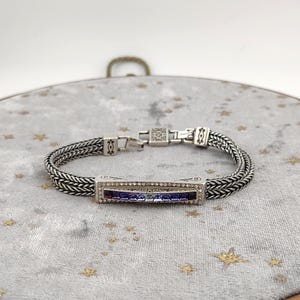 K&ouml;nnte beinhalten: Silberfarbenes Armband mit einem rechteckigen Mittelst&uuml;ck mit einem Farbverlauf aus lila und blauen Steinen. Das Armband hat ein geflochtenes Kettenmuster und einen sicheren Verschluss. Das Mittelst&uuml;ck ist mit klaren Kristallen umrahmt.