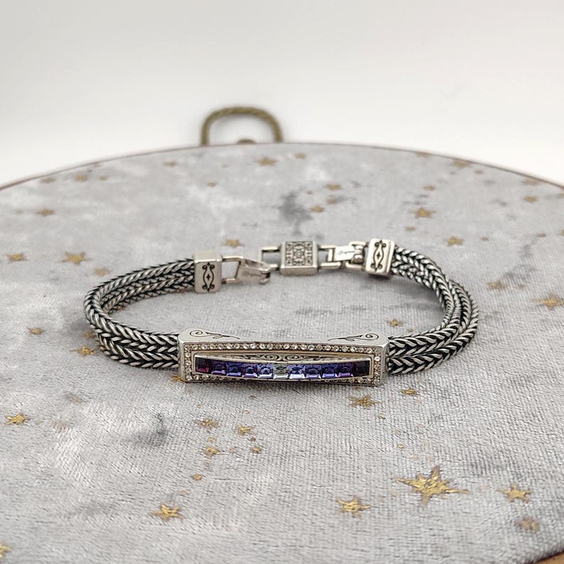 K&ouml;nnte beinhalten: Silberfarbenes Armband mit einem rechteckigen Mittelst&uuml;ck mit einem Farbverlauf aus lila und blauen Steinen. Das Armband hat ein geflochtenes Kettenmuster und einen sicheren Verschluss. Das Mittelst&uuml;ck ist mit klaren Kristallen umrahmt.