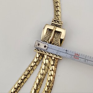 May include: Gold-toned vintage belt with a buckle design. The belt features three chain strands. The buckle is approximately 5 cm wide. The belt is measured with a tape measure.