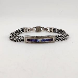 K&ouml;nnte beinhalten: Silberfarbenes Armband mit geflochtenem Kettenmuster. Das Armband hat einen rechteckigen Mittelteil mit einer Reihe kleiner, klarer Steine und einer Reihe blauer Steine. Der Verschluss hat ein quadratisches Design mit dekorativen Elementen.