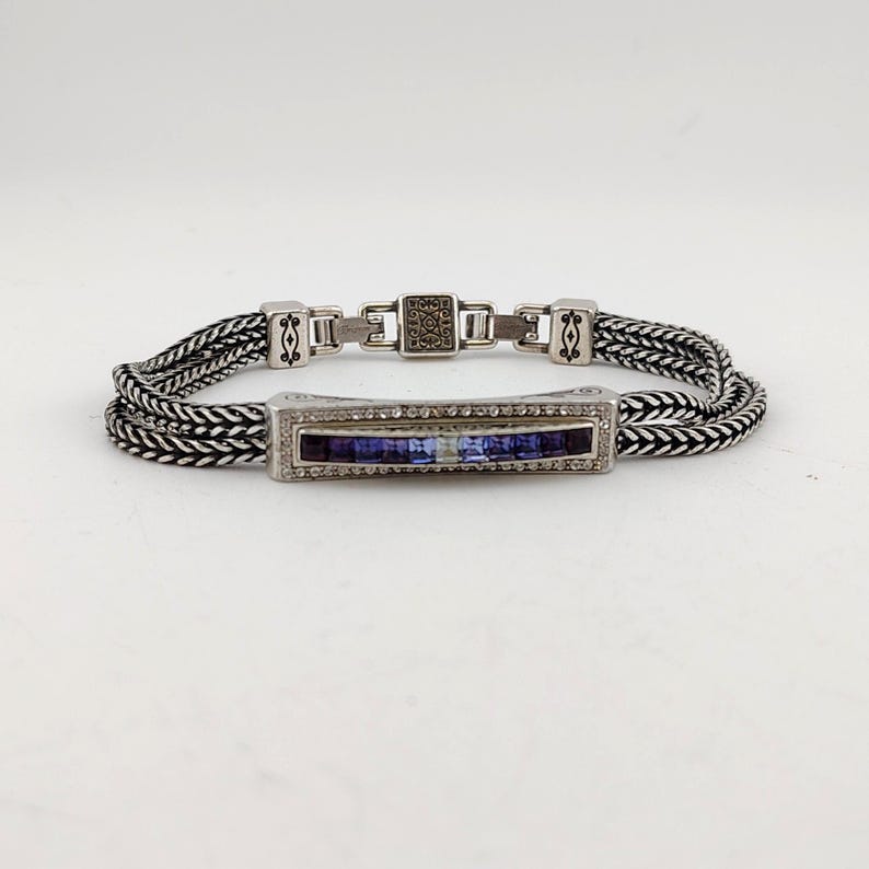 K&ouml;nnte beinhalten: Silberfarbenes Armband mit geflochtenem Kettenmuster. Das Armband hat einen rechteckigen Mittelteil mit einer Reihe kleiner, klarer Steine und einer Reihe blauer Steine. Der Verschluss hat ein quadratisches Design mit dekorativen Elementen.