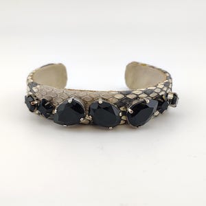 May include: A cuff bracelet featuring a snakeskin pattern in shades of gray and black. The bracelet is adorned with several large, black teardrop-shaped gemstones set in silver-toned metal. The bracelet has an open design.