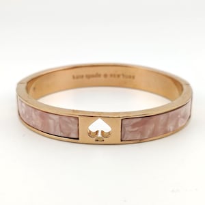 May include: A rose gold-tone hinged bangle bracelet featuring a central spade-shaped cutout and two inlaid panels of light pink marbled resin. The interior of the metal band is engraved with the text kate spade new york in a sans-serif font.