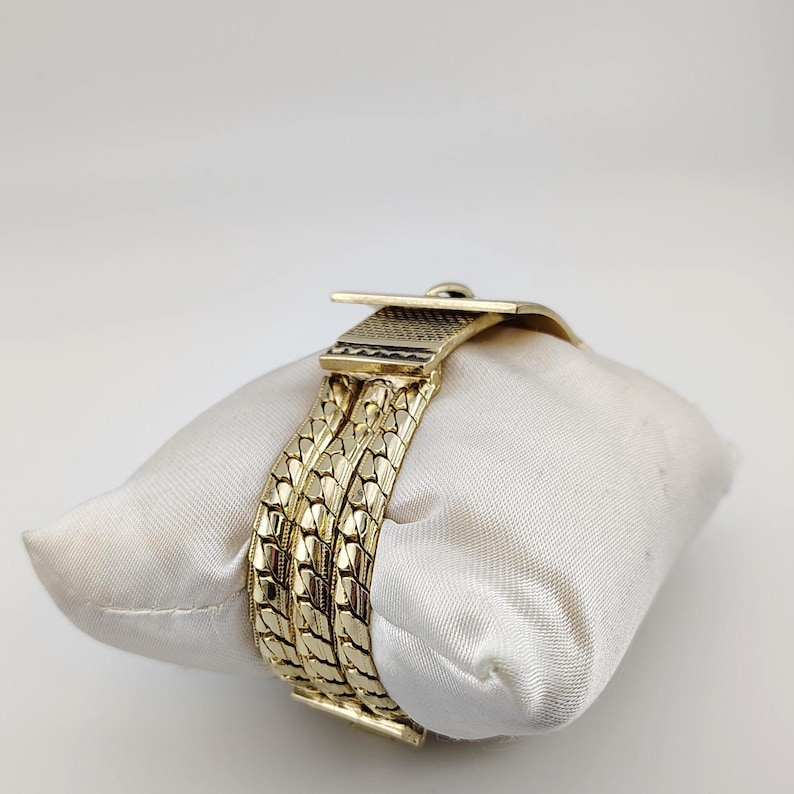 May include: A gold bracelet with a woven pattern and a buckle clasp. The bracelet is presented on a white satin cushion, highlighting its detailed design and polished surface. The bracelet's design is made up of three parallel bands.