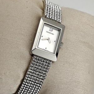 Può includere: Un orologio da polso rettangolare color argento con quadrante bianco che riporta il logo del cigno Swarovski e la scritta. L'orologio presenta un cinturino metallico densamente incastonato di piccoli cristalli chiari e scintillanti.