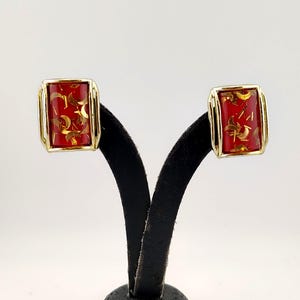 May include: A pair of vintage rectangular clip-on earrings. Each earring features a red center with gold-colored abstract shapes, framed by a gold-tone metal border. The earrings are displayed on a black stand against a white background.