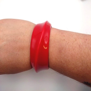 May include: A vibrant red bangle bracelet with a curved design. The bracelet has a smooth, glossy finish and a small, gold-colored accent. This bold accessory is a statement piece.