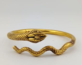 May include: A gold-coloured snake bracelet. The bracelet features a detailed snake head and body, with scales and a curved design. The snake's head is at one end, and the body curves around to form the bracelet.