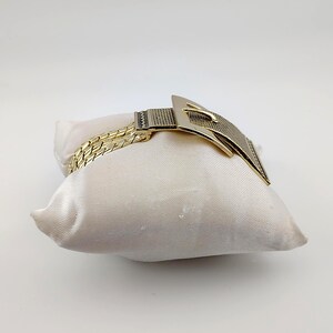 May include: A gold-coloured bracelet with a buckle design, displayed on a cream satin pillow. The bracelet has a woven chain and a textured buckle. The buckle is rectangular with a pointed end.