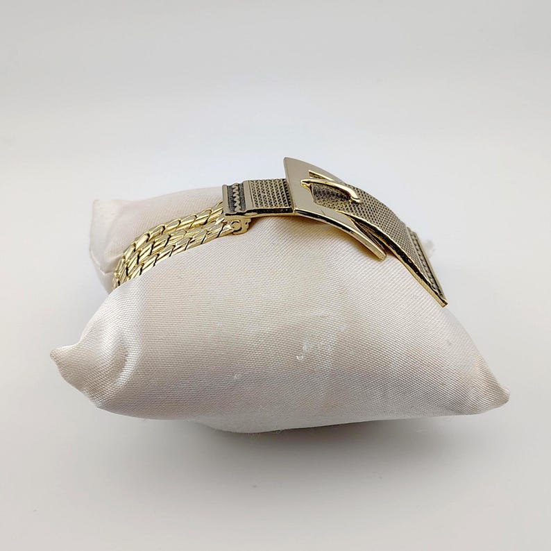 May include: A gold-coloured bracelet with a buckle design, displayed on a cream satin pillow. The bracelet has a woven chain and a textured buckle. The buckle is rectangular with a pointed end.