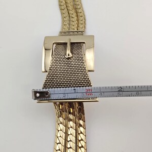 May include: Gold-coloured belt featuring a large rectangular buckle with a textured mesh design and a classic pin closure. The belt's chain is measured with a tape measure, showing a width of approximately 5 cm.