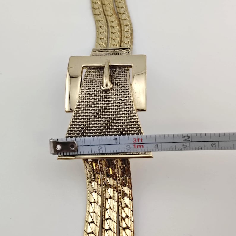 May include: Gold-coloured belt featuring a large rectangular buckle with a textured mesh design and a classic pin closure. The belt's chain is measured with a tape measure, showing a width of approximately 5 cm.