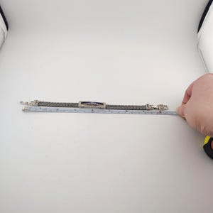 K&ouml;nnte beinhalten: Silberfarbenes Armband mit geflochtenem Kettenmuster. Das Armband hat ein rechteckiges Mittelst&uuml;ck mit blauen und wei&szlig;en Akzenten, umgeben von kleinen, klaren Steinen. Das Armband ist ca. 17,8 cm lang.