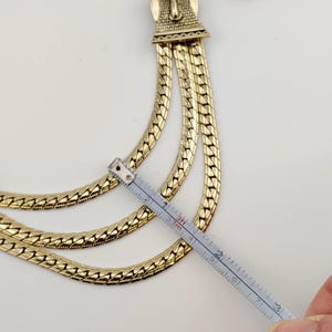 May include: Gold-coloured necklace with three layered chains. The necklace is measured with a ruler, showing a length of approximately 7.6 cm. The clasp has an ornate design. The background is white.