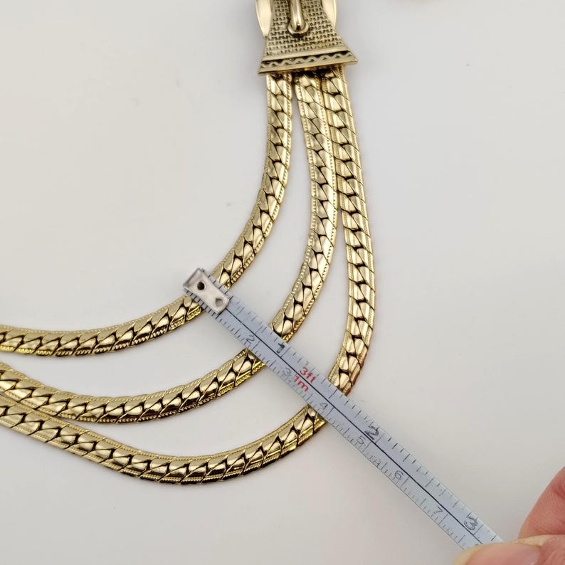 May include: Gold-coloured necklace with three layered chains. The necklace is measured with a ruler, showing a length of approximately 7.6 cm. The clasp has an ornate design. The background is white.