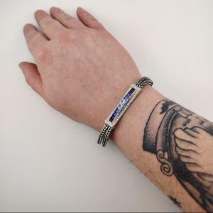 K&ouml;nnte beinhalten: Silbernes Armband mit einem rechteckigen Mittelteil, verziert mit lila und blauen Edelsteinen und kleinen klaren Steinen. Das Armband hat ein geflochtenes Design und wird am Handgelenk getragen.
