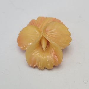 Könnte beinhalten: Eine Brosche in Form einer geschnitzten Blume in blassgelber und rosa Farbe. Die Blume hat fünf Blütenblätter und eine Mitte mit einer kleinen, spitzen Knospe.
