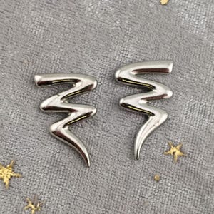 May include: A pair of silver-colored earrings in a zigzag design. The earrings have a polished finish and a curved, elongated shape. The earrings are displayed on a gray surface with small gold star-shaped decorations.