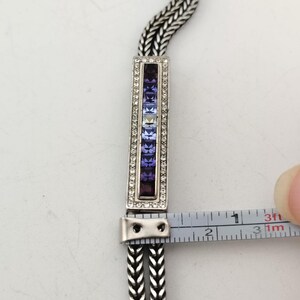 K&ouml;nnte beinhalten: Silberfarbenes Armband mit rechteckigem Mittelteil. Das Mittelteil ist mit einem Farbverlauf aus lila und klaren Edelsteinen verziert. Das Armband wird mit einem Lineal gemessen, ca. 2,5 cm lang.