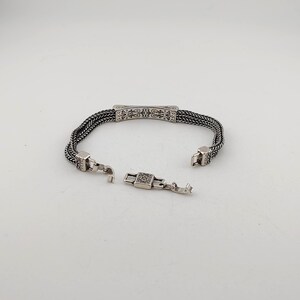 K&ouml;nnte beinhalten: Silberarmband mit offenem Verschluss. Das Armband hat ein geflochtenes Kettendesign mit einem dekorativen rechteckigen Mittelteil und quadratischen Verschlussenden. Der Verschluss ist ge&ouml;ffnet und zeigt die komplizierten Details des Schmucks.