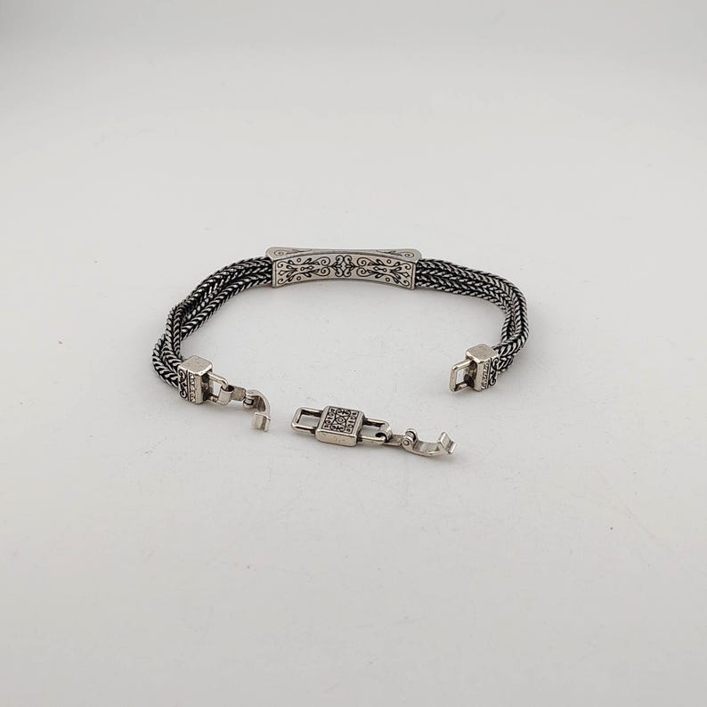K&ouml;nnte beinhalten: Silberarmband mit offenem Verschluss. Das Armband hat ein geflochtenes Kettendesign mit einem dekorativen rechteckigen Mittelteil und quadratischen Verschlussenden. Der Verschluss ist ge&ouml;ffnet und zeigt die komplizierten Details des Schmucks.