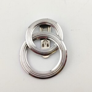 May include: A silver-toned metal scarf clip featuring two interlocking circular rings with a grooved, concentric design. The back of the accessory includes a flat, vertical metal clasp mechanism with a small engraved leaf-like detail at the top.