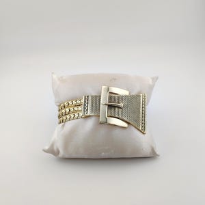 May include: Gold-coloured bracelet with a buckle design. The bracelet features a woven band and a rectangular buckle with a textured surface. The bracelet is displayed on a cream-coloured pillow.