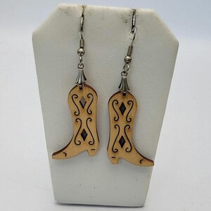May include: A pair of wooden cowboy boot earrings. The earrings are light brown with black etched details and silver-colored ear wires. The boots have decorative cutouts and scrollwork.