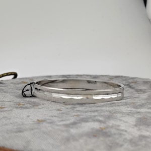 May include: A silver bangle bracelet with a textured design. The bracelet has a smooth, polished finish with a series of horizontal lines and a textured pattern. The bracelet is displayed on a gray surface.
