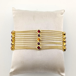 May include: A gold-toned multi-strand bracelet featuring seven thin, flexible snake chains. The center of the bracelet is adorned with a vertical row of five alternating square and round faceted crystals in shades of red, purple, and orange.