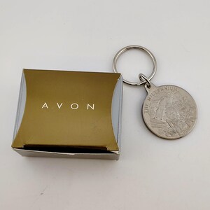 May include: A gold-colored box with the word "AVON" in white lettering sits next to a silver-colored keychain. The keychain has a round pendant with an engraved design and the words "THE MILLENNIUM" and "2000".