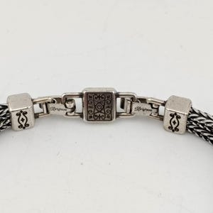 K&ouml;nnte beinhalten: Nahaufnahme eines silberfarbenen Armbands mit einer gewebten schwarz-silbernen Kette. Das Armband hat einen quadratischen Verschluss mit einem verzierten Design und quadratischen Endst&uuml;cken mit dekorativen Elementen. Das Wort "Brighton" ist auf dem Verschluss zu sehen.