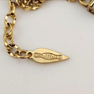May include: Close-up of a gold-toned necklace chain with a leaf-shaped tag. The tag is engraved with the text "Whiting & Davis Co." The chain is made of interlocking links and is set against a white background.