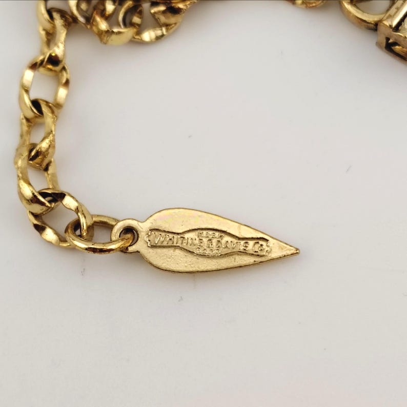 May include: Close-up of a gold-toned necklace chain with a leaf-shaped tag. The tag is engraved with the text "Whiting & Davis Co." The chain is made of interlocking links and is set against a white background.