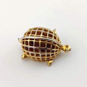 May include: A gold-toned turtle brooch with a silver band encircling its body. The turtle's shell is a grid pattern, and it has small, detailed feet and a head with tiny eyes. The brooch is set against a white background.
