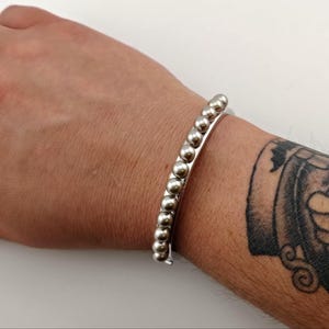 May include: A silver-toned cuff bracelet featuring a single row of smooth, spherical beads arranged along the outer edge. The bracelet has a polished metallic finish and a curved, open-ended design that rests securely around the wrist area.
