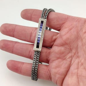 K&ouml;nnte beinhalten: Silberarmband mit rechteckigem Mittelteil, das einen Farbverlauf aus blauen und violetten Edelsteinen aufweist, umrandet von kleinen, klaren Steinen. Das Armband hat ein gewebtes Kettenmuster.