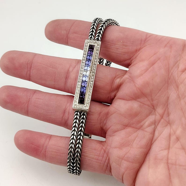 K&ouml;nnte beinhalten: Silberarmband mit rechteckigem Mittelteil, das einen Farbverlauf aus blauen und violetten Edelsteinen aufweist, umrandet von kleinen, klaren Steinen. Das Armband hat ein gewebtes Kettenmuster.