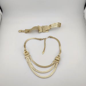 May include: A gold-coloured necklace with three layered chains and a matching belt. The necklace has decorative rectangular clasps on each side. The belt features a rectangular buckle. Both pieces are made of a woven metal material.
