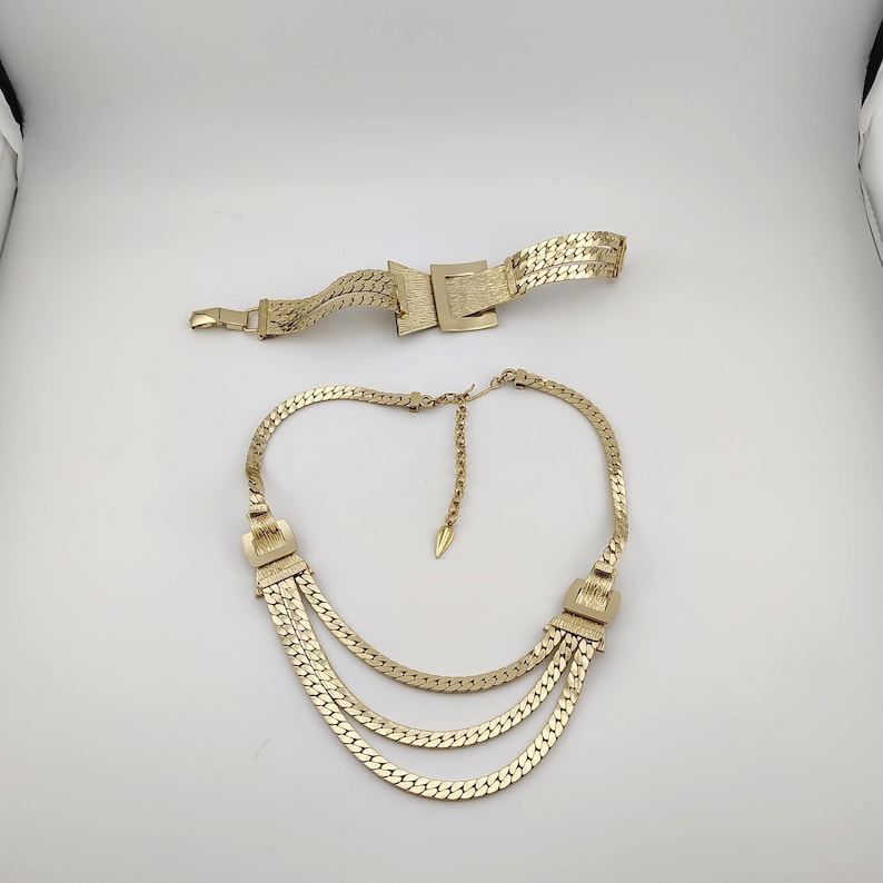 May include: A gold-coloured necklace with three layered chains and a matching belt. The necklace has decorative rectangular clasps on each side. The belt features a rectangular buckle. Both pieces are made of a woven metal material.