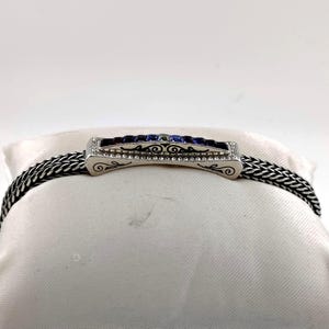 K&ouml;nnte beinhalten: Silberfarbenes Armband mit geflochtener Kette und rechteckigem Mittelst&uuml;ck. Das Mittelst&uuml;ck hat blaue und violette Edelsteine, kleine klare Steine und dekorative Schn&ouml;rkel. Das Armband wird auf einer wei&szlig;en Oberfl&auml;che pr&auml;sentiert.