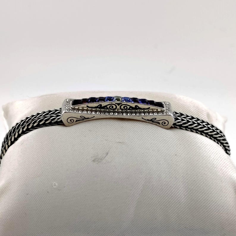 K&ouml;nnte beinhalten: Silberfarbenes Armband mit geflochtener Kette und rechteckigem Mittelst&uuml;ck. Das Mittelst&uuml;ck hat blaue und violette Edelsteine, kleine klare Steine und dekorative Schn&ouml;rkel. Das Armband wird auf einer wei&szlig;en Oberfl&auml;che pr&auml;sentiert.