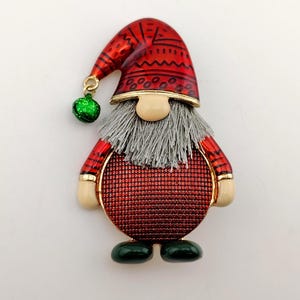May include: A gnome-shaped brooch featuring a red hat with black geometric patterns and a dangling green bell. The figure has a textured grey beard, a round beige nose, a red glittery body, red sleeves with black stripes, and dark green shoes.