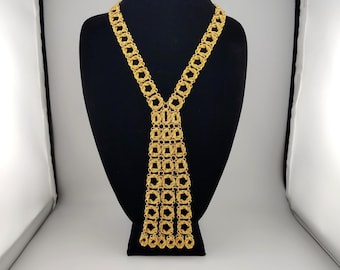 May include: A gold-toned necklace featuring a long, tie-like design constructed from interconnected circular metal links. The piece consists of two strands that converge into a wider, rectangular panel of woven chain links that hangs vertically.