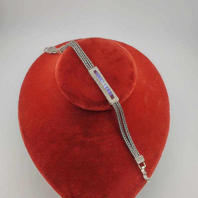 K&ouml;nnte beinhalten: Ein silbernes Armband mit einem rechteckigen Mittelst&uuml;ck mit einer Reihe kleiner, klarer Steine und einem Farbverlauf aus lila und blauen Steinen. Das Armband hat ein gewebtes Kettendesign und wird auf einer roten Samtoberfl&auml;che pr&auml;sentiert.