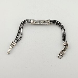 K&ouml;nnte beinhalten: Silberfarbenes Armband mit geflochtenem Kettenmuster. Das Armband hat ein verziertes rechteckiges Mittelst&uuml;ck mit dekorativen Mustern. Der Verschluss und die Verbindungselemente sind ebenfalls silberfarben, was einen stimmigen Look erzeugt. Das Armband ist ca. 17,78 cm lang.