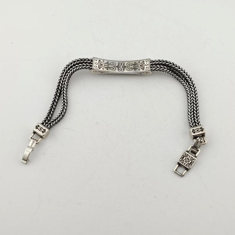 K&ouml;nnte beinhalten: Silberfarbenes Armband mit geflochtenem Kettenmuster. Das Armband hat ein verziertes rechteckiges Mittelst&uuml;ck mit dekorativen Mustern. Der Verschluss und die Verbindungselemente sind ebenfalls silberfarben, was einen stimmigen Look erzeugt. Das Armband ist ca. 17,78 cm lang.